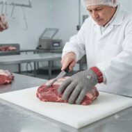 Butcher in a white coat and hairnet slices a large piece of raw beef on a white cutting board with a knife, in a sterile kitchen.