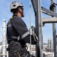 Industrial worker in helmet and safety harness adjusting a steel cable on a metal scaffold at an industrial site.