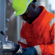 Industrial worker in orange safety coveralls and a neon yellow hard hat inspecting a metal valve fitting. Use of a tool is implied near the connector.