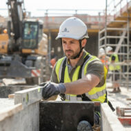 Construction worker in a yellow safety vest and white hard hat places a wooden plank on a concrete form at a busy site.