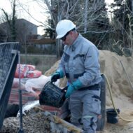 Construction worker in a hard hat and gloves pouring material from a bucket into a cement mixer at a construction site that has sand piles and bags nearby.