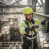 Construction worker in a yellow helmet and high-visibility vest tightening a bolt on a steel beam at a construction site with scaffolding in the background.