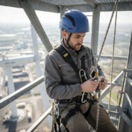 Construction worker wearing a blue hard hat and safety harness on a high metal structure, adjusting climbing ropes.