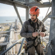 Construction worker in an orange helmet and safety harness on a high steel platform overlooking an industrial facility by the sea.