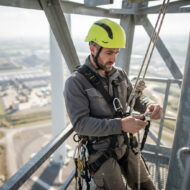 Worker wearing a yellow helmet and safety harness adjusts ropes on a high metal scaffold, with a distant cityscape below.