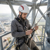 Construction worker in a white helmet and gray coveralls securing rope to a carabiner on a tall steel tower, high above the ground.