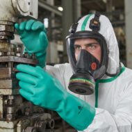 Worker in a white hazmat suit and teal gloves operates a rusty industrial machine with a lever, wearing a full-face respirator for protection