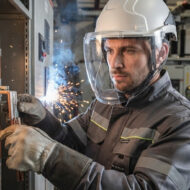 Male industrial welder in a protective helmet and face shield welding metal, sparks flying in a workshop setting.