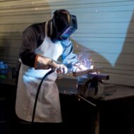 Welder in protective gear welding metal on a workbench, sparks flying around.