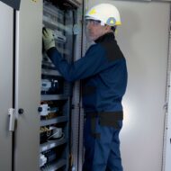 Technician in safety gear inspecting an open electrical cabinet with wiring and controls