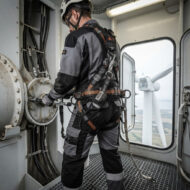 Maintenance worker in a full-body safety harness inspecting large machinery inside a wind turbine nacelle, with the rotor seen through the open door