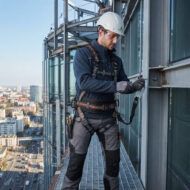 Construction worker on a high-rise exterior wearing helmet and full-body harness, securing equipment on a metal ledge with cityscape below