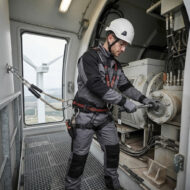 Maintenance technician in a hard hat and safety harness tightening a large gearbox inside a wind turbine nacelle, with the turbine visible through the window