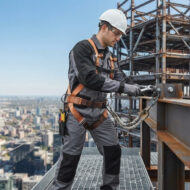 Construction worker in a hard hat and safety harness on a high-rise steel scaffold, tightening a rope with a wrench while overlooking the city.