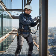 Construction worker wearing a hard hat and safety harness attaches a bolt to a steel beam on the exterior of a high-rise building.