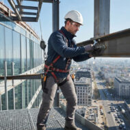 Construction worker in a hard hat and safety harness tightens a bolt on a steel beam high above a city skyline.