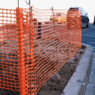 Orange plastic construction fencing along a curb, marking a restricted construction area by a road with passing cars in the background.