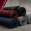 Rolled exercise mats in blue, red, black, and gray stacked on a gym floor with a concrete backdrop and maroon diagonal stripes (informative).
