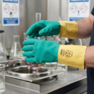 Scientist wearing green and yellow protective gloves pours a liquid into a beaker in a laboratory setup with safety signs in the background