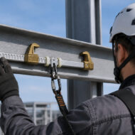 Construction worker in a white safety helmet adjusting a metal beam clamp with a carabiner and strap.
