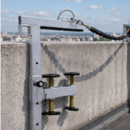 Rooftop anchor device with bolts and a cable-connected safety lanyard over a concrete parapet, used for fall protection