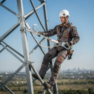 Worker in helmet and safety harness on a steel lattice tower, using a tool while secured with a rope.