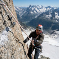 Climber wearing helmet and harness ascends a rock face, secured by rope and carabiners with a snowy mountain valley in the background