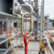 Gold carabiner clipped to a metal anchor on a construction scaffold with a red rope tether.