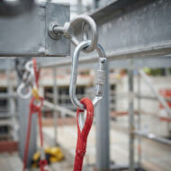 Close-up of a metal carabiner clipped to a circular steel link on a construction scaffold, with a red rope threaded through.
