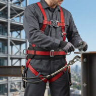Construction worker wearing a red-and-black full-body safety harness with lanyard, standing on a steel beam at a construction site.