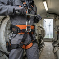 Worker in gray coveralls wearing full-body orange waist harness with carabiners and tools, standing in an industrial maintenance cabin