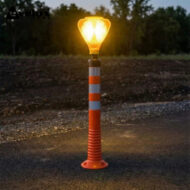 Illuminated amber safety beacon on an orange-and-white striped post at a construction site, glowing in dusk.