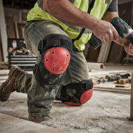 Construction worker kneeling on a dusty floor wearing a neon safety vest and red knee pads, using a cordless drill.