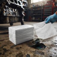 Gloved hand uses a paper towel to wipe a dark oil spill on a workshop floor, with an engine block in the background.