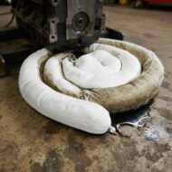 Coiled white fabric insulation sleeve placed under a heavy machine in a workshop, with an oil puddle nearby on the floor left blank.