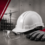 White construction hard hat, clear safety goggles, and red gloves on a work surface with a building site and crane in the background.